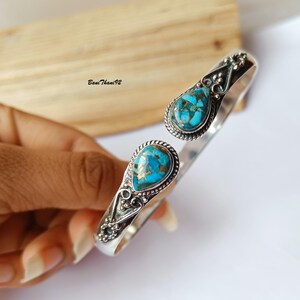 Può includere: Bracciale aperto con pietre a goccia turchesi e color rame incastonate in metallo argentato. Il bracciale ha un design ornato con dettagli decorativi attorno alle pietre e lungo la fascia.