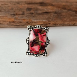 Può includere: Anello color argento con pietra rettangolare composita. La pietra presenta una miscela di tonalità rosse, rosa e oro. L'anello ha un bordo decorativo con perline, su sfondo neutro.