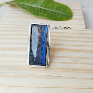 May include: A rectangular silver ring featuring a large, polished labradorite stone. The stone displays a mix of blue, gray, and black hues. The ring is set on a light wooden surface, with a green leaf in the background.
