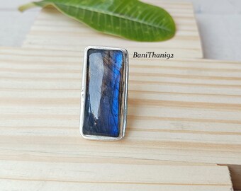 Può includere: Un anello rettangolare in argento con una pietra di labradorite lucidata. La pietra mostra una miscela di tonalità blu, grigie e nere. L'anello è posto su una superficie di legno chiaro, con una foglia verde sullo sfondo.