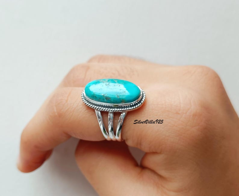 May include: A silver ring featuring a large, oval turquoise stone. The stone is a vibrant blue-green color, set in a detailed silver band with a braided edge. The ring has a split shank design.