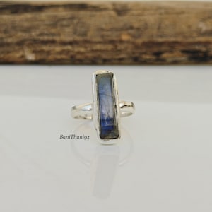 May include: A silver ring with a rectangular, iridescent blue gemstone. The ring has a simple band and a bezel setting. The gemstone displays shades of blue and gray. The ring is on a white surface.
