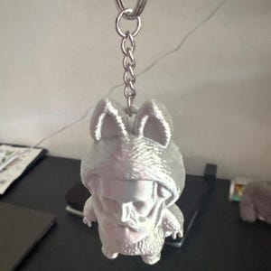 May include: A silver-colored keychain featuring a 3D-printed figure of a creature wearing a hooded outfit. The figure has a skull-like face and bunny ears. The keychain includes a metal ring and chain. The word "SWM" is visible in the background.