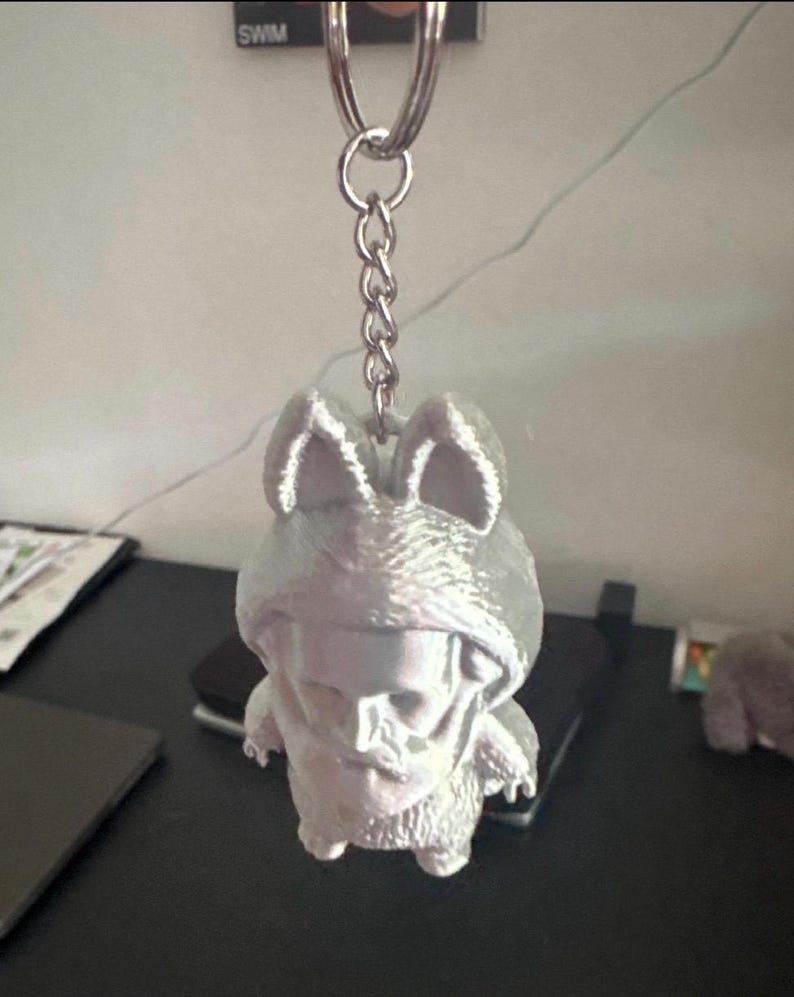 May include: A silver-colored keychain featuring a 3D-printed figure of a creature wearing a hooded outfit. The figure has a skull-like face and bunny ears. The keychain includes a metal ring and chain. The word "SWM" is visible in the background.