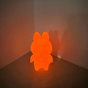May include: An illuminated orange figurine of a cartoon character with a rounded body, small arms, and large ears. The figure is glowing brightly, casting a shadow on the wall behind it. The product is a night light.
