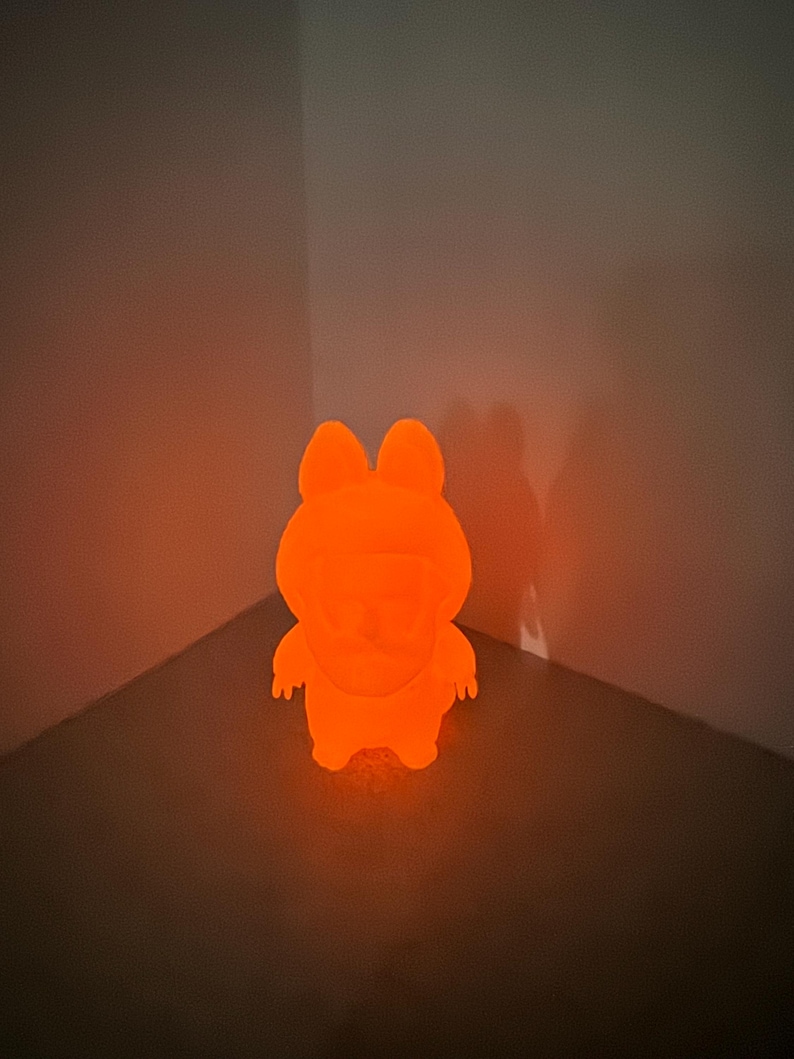 May include: An illuminated orange figurine of a cartoon character with a rounded body, small arms, and large ears. The figure is glowing brightly, casting a shadow on the wall behind it. The product is a night light.