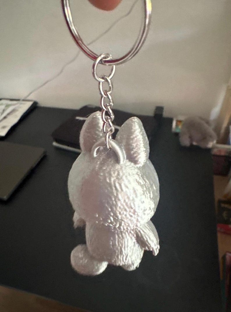 May include: A silver-colored bunny keychain with a textured surface. The bunny is attached to a silver chain and keyring. The keychain is a cute accessory.