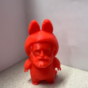 Könnte beinhalten: Eine leuchtend rote Figur mit einem cartoonartigen Design. Die Figur hat ein Gesicht, das einer Person ähnelt, mit großen Ohren und einem kleinen Körper. Die Figur besteht aus einem festen Material und steht auf einer gesprenkelten Oberfläche.