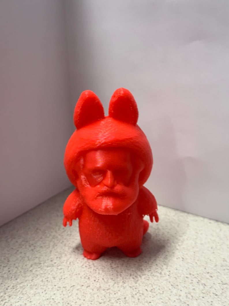 May include: A bright red figurine with a cartoonish design. The figure has a face resembling a person, with large ears and a small body. The figurine is made of a solid material and stands on a speckled surface.