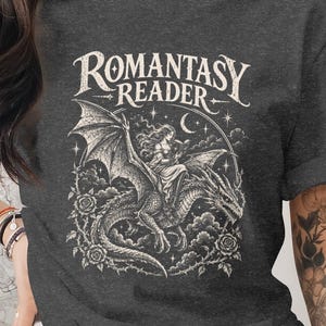 Può includere: Una t-shirt grigio antracite mélange con una stampa grafica bianca che raffigura una persona a cavallo di un drago circondato da rose, stelle e una luna crescente. Il testo ROMANTASY READER è stampato in un font stilizzato sopra l'immagine.