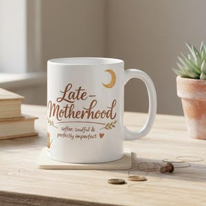 May include: A white ceramic mug with the words "Late Motherhood" in brown script, along with the phrases "softer, soulful & perfectly imperfect." A crescent moon and leaf accents are also present. The mug sits on a wooden surface.