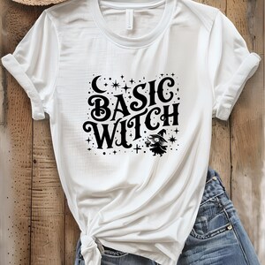 May include: White t-shirt with the text "BASIC WITCH" in a black, stylized font. The design includes a crescent moon, stars, and a small witch illustration holding a potion. The shirt is folded and placed on a wooden surface.