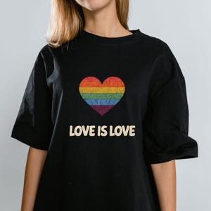 Puede incluir: Camiseta negra con un diseño de corazón arcoíris. El corazón está compuesto por rayas horizontales de varios colores. Debajo del corazón, el texto "LOVE IS LOVE" está impreso en un estilo vintage. La camiseta tiene mangas cortas.