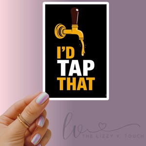 Op de afbeelding: Een sticker met een zwarte achtergrond en de tekst "I'd TAP THAT" in wit en geel. Boven de tekst staat een goudkleurige biertap. De sticker wordt vastgehouden door een hand met iriserende nagels.