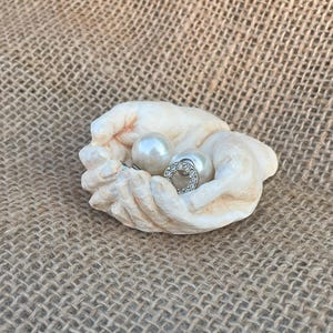 Puede incluir: Un plato en forma de manos esculpidas de color crema que contiene dos pendientes de perlas. Uno es una perla grande y redonda, y el otro presenta una perla más pequeña rodeada por un diseño circular plateado con piedras transparentes.