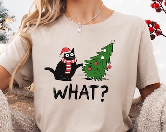 May include: Beige t-shirt featuring a black cat wearing a Santa hat and a red and white striped scarf, next to a decorated Christmas tree. The word "WHAT?" is printed below the cat and tree. The shirt is part of a holiday collection.