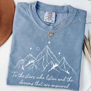 Puede incluir: Camiseta azul vaquero Comfort Colors con un gr&aacute;fico de monta&ntilde;a y el texto "To the stars who listen and the dreams that are answered." Un collar dorado con un colgante est&aacute; sobre la camiseta.