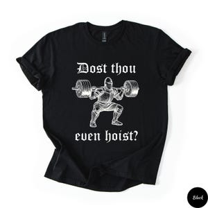 Op de afbeelding: Zwart T-shirt met een witte afbeelding van een ridder die een halter heft. De tekst "Dost thou even hoist?" staat erboven en eronder. Het shirt heeft opgerolde mouwen en een ronde hals.