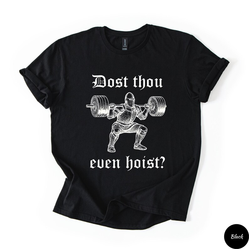 Op de afbeelding: Zwart T-shirt met een witte afbeelding van een ridder die een halter heft. De tekst "Dost thou even hoist?" staat erboven en eronder. Het shirt heeft opgerolde mouwen en een ronde hals.