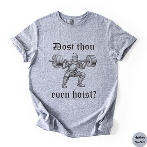 Op de afbeelding: Atletisch heather grijze t-shirt met een zwarte afbeelding van een ridder die gewichten heft, met de tekst "Dost thou even hoist?" in een middeleeuws lettertype. Het shirt is gemaakt van een zacht, comfortabel materiaal.