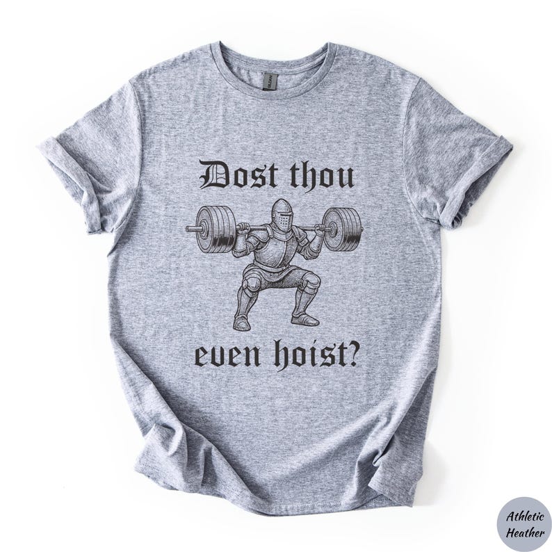Op de afbeelding: Atletisch heather grijze t-shirt met een zwarte afbeelding van een ridder die gewichten heft, met de tekst "Dost thou even hoist?" in een middeleeuws lettertype. Het shirt is gemaakt van een zacht, comfortabel materiaal.