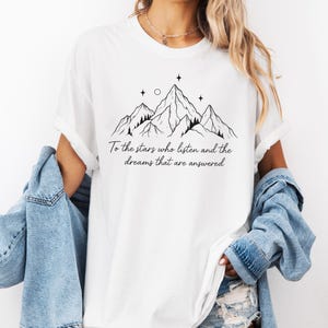 Puede incluir: Camiseta blanca con un dise&ntilde;o de cordillera negra, estrellas y el texto "To the stars who listen and the dreams that are answered." La camiseta tiene cuello redondo y mangas cortas.