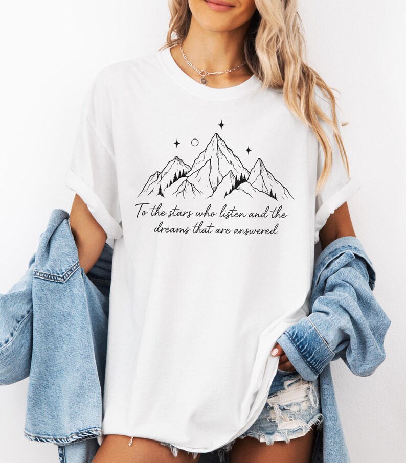 Puede incluir: Camiseta blanca con un dise&ntilde;o de cordillera negra, estrellas y el texto "To the stars who listen and the dreams that are answered." La camiseta tiene cuello redondo y mangas cortas.