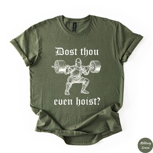 Op de afbeelding: Militairgroen t-shirt met opgerolde mouwen. Het shirt heeft een witte afbeelding van een ridder die gewichten heft, met de tekst "Dost thou even hoist?" in een middeleeuws lettertype. Het shirt is gemaakt van zacht materiaal.