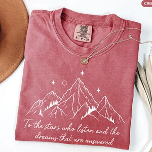 Puede incluir: Camiseta de color carmes&iacute; con un dise&ntilde;o de monta&ntilde;a blanco y el texto "To the stars who listen and the dreams that are answered." La camiseta tiene cuello redondo y mangas cortas. Un collar plateado con un colgante est&aacute; sobre la camiseta.