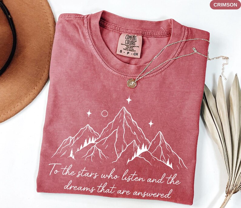 Puede incluir: Camiseta de color carmes&iacute; con un dise&ntilde;o de monta&ntilde;a blanco y el texto "To the stars who listen and the dreams that are answered." La camiseta tiene cuello redondo y mangas cortas. Un collar plateado con un colgante est&aacute; sobre la camiseta.
