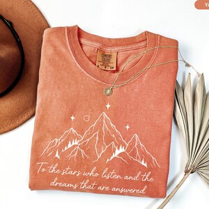 Puede incluir: Una camiseta de color coral con un gr&aacute;fico de monta&ntilde;a y el texto "To the stars who listen and the dreams that are answered." Tambi&eacute;n se ven un collar dorado y un sombrero marr&oacute;n.