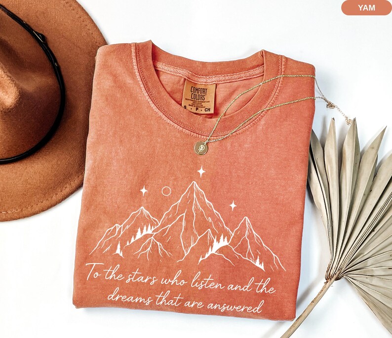 Puede incluir: Una camiseta de color coral con un gr&aacute;fico de monta&ntilde;a y el texto "To the stars who listen and the dreams that are answered." Tambi&eacute;n se ven un collar dorado y un sombrero marr&oacute;n.