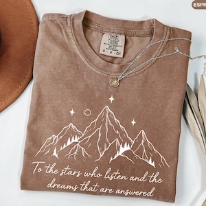 Puede incluir: Camiseta color espresso con un gr&aacute;fico de monta&ntilde;a blanco y el texto "To the stars who listen and the dreams that are answered." La camiseta tiene cuello redondo y una etiqueta Comfort Colors. Un collar plateado con un colgante est&aacute; sobre la camiseta.