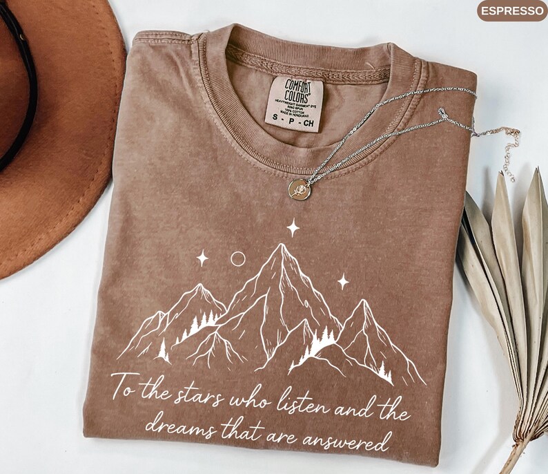Puede incluir: Camiseta color espresso con un gr&aacute;fico de monta&ntilde;a blanco y el texto "To the stars who listen and the dreams that are answered." La camiseta tiene cuello redondo y una etiqueta Comfort Colors. Un collar plateado con un colgante est&aacute; sobre la camiseta.