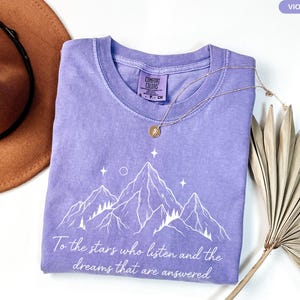 Puede incluir: Una camiseta violeta Comfort Colors con un gr&aacute;fico de monta&ntilde;a y el texto "To the stars who listen and the dreams that are answered." Un collar dorado y un sombrero marr&oacute;n tambi&eacute;n est&aacute;n en la imagen.