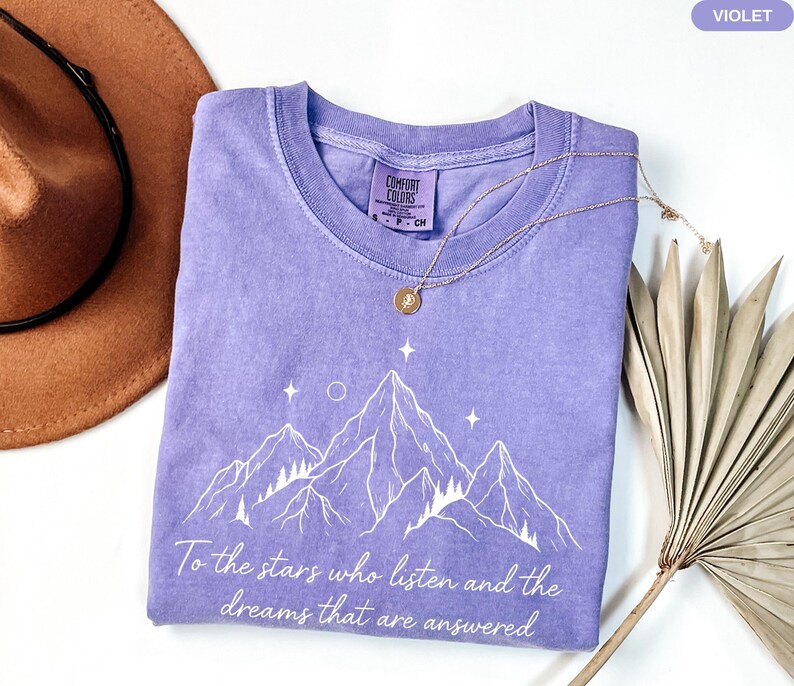 Puede incluir: Una camiseta violeta Comfort Colors con un gr&aacute;fico de monta&ntilde;a y el texto "To the stars who listen and the dreams that are answered." Un collar dorado y un sombrero marr&oacute;n tambi&eacute;n est&aacute;n en la imagen.