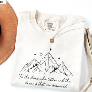 Puede incluir: Camiseta Comfort Colors color marfil con un dise&ntilde;o de monta&ntilde;a y el texto "To the stars who listen and the dreams that are answered." Un collar dorado con un colgante est&aacute; sobre la camiseta.