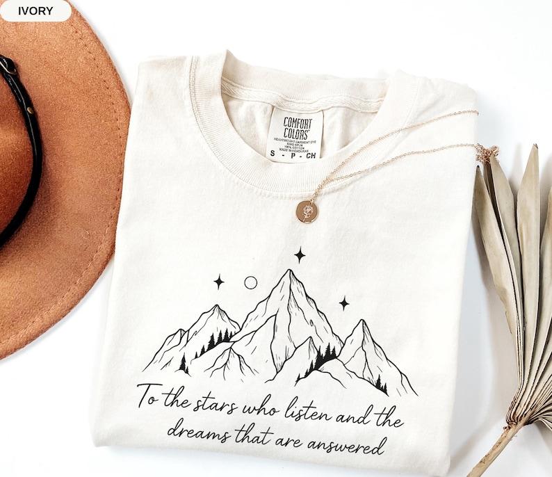 Puede incluir: Camiseta Comfort Colors color marfil con un dise&ntilde;o de monta&ntilde;a y el texto "To the stars who listen and the dreams that are answered." Un collar dorado con un colgante est&aacute; sobre la camiseta.