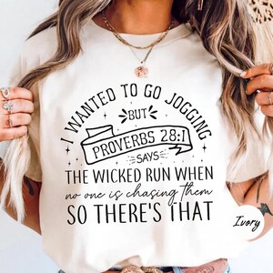 May include: Ivory-colored t-shirt with black text that reads "I WANTED TO GO JOGGING BUT PROVERBS 28:1 SAYS THE WICKED RUN WHEN no one is chasing them SO THERE'S THAT." The shirt has a crew neck.