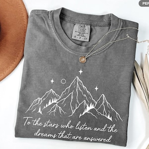 Puede incluir: Camiseta gris con un gr&aacute;fico de monta&ntilde;a y el texto "To the stars who listen and the dreams that are answered." La camiseta tiene cuello redondo y mangas cortas. Un collar plateado con un colgante est&aacute; sobre la camiseta.