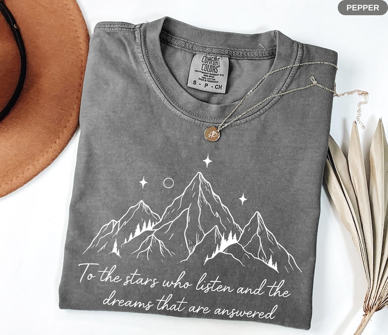 Puede incluir: Camiseta gris con un gr&aacute;fico de monta&ntilde;a y el texto "To the stars who listen and the dreams that are answered." La camiseta tiene cuello redondo y mangas cortas. Un collar plateado con un colgante est&aacute; sobre la camiseta.