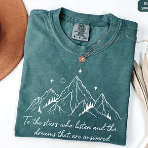 Puede incluir: Camiseta azul abeto con un gr&aacute;fico de monta&ntilde;a y el texto "To the stars who listen and the dreams that are answered". La camiseta tiene cuello redondo y mangas cortas. Un collar dorado con un colgante est&aacute; sobre la camiseta.