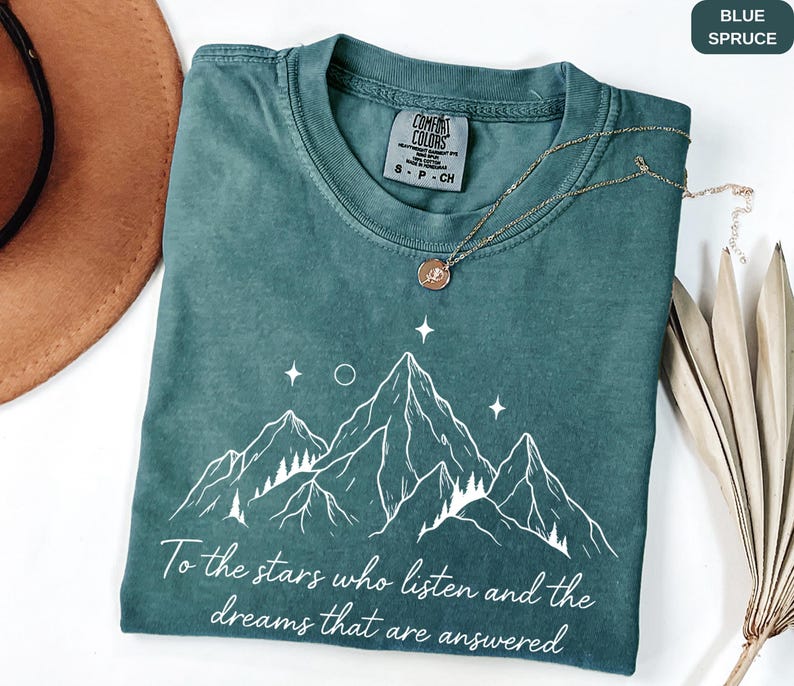 Puede incluir: Camiseta azul abeto con un gr&aacute;fico de monta&ntilde;a y el texto "To the stars who listen and the dreams that are answered". La camiseta tiene cuello redondo y mangas cortas. Un collar dorado con un colgante est&aacute; sobre la camiseta.