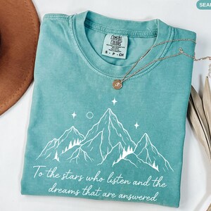 Puede incluir: Camiseta verde mar con un gr&aacute;fico de monta&ntilde;a y el texto "To the stars who listen and the dreams that are answered." La camiseta tiene cuello redondo y una peque&ntilde;a etiqueta que dice "COMFORT COLORS". Un collar dorado est&aacute; drapeado sobre la camiseta.