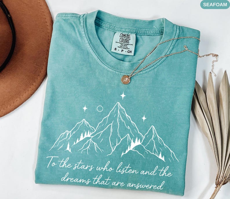 Puede incluir: Camiseta verde mar con un gr&aacute;fico de monta&ntilde;a y el texto "To the stars who listen and the dreams that are answered." La camiseta tiene cuello redondo y una peque&ntilde;a etiqueta que dice "COMFORT COLORS". Un collar dorado est&aacute; drapeado sobre la camiseta.