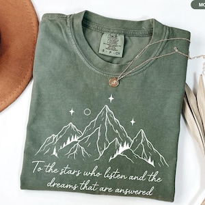 Puede incluir: Camiseta verde musgo con un dise&ntilde;o de monta&ntilde;a blanca y el texto "To the stars who listen and the dreams that are answered." Un collar dorado con un colgante descansa sobre la camiseta. La camiseta es de material suave.