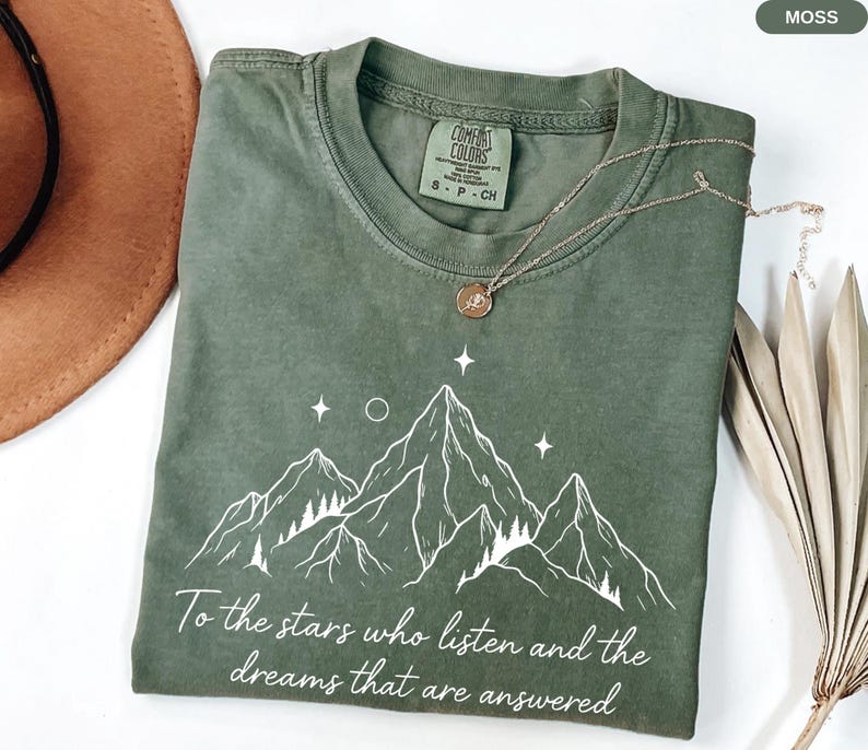 Puede incluir: Camiseta verde musgo con un dise&ntilde;o de monta&ntilde;a blanca y el texto "To the stars who listen and the dreams that are answered." Un collar dorado con un colgante descansa sobre la camiseta. La camiseta es de material suave.