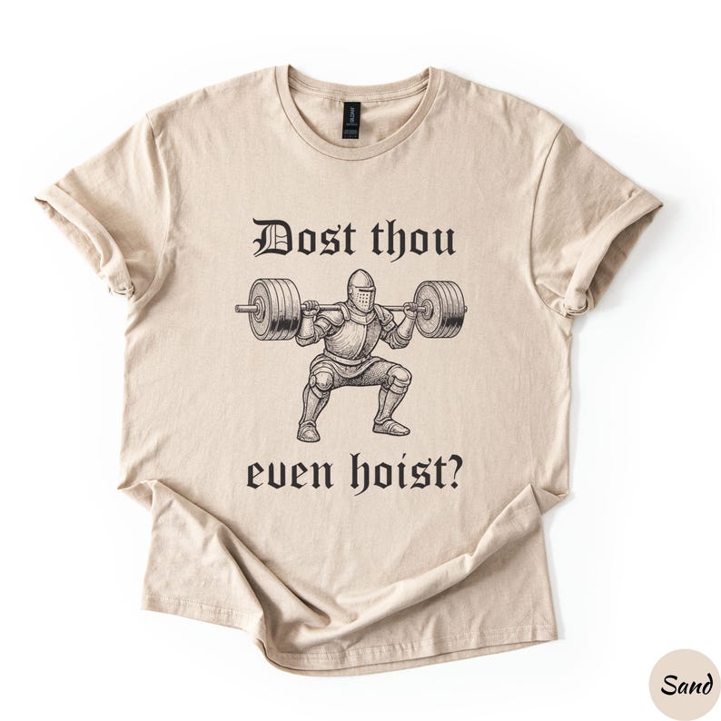 Op de afbeelding: Zandkleurig T-shirt met een zwarte afbeelding van een ridder die gewichten heft, en de tekst "Dost thou even hoist?" in een middeleeuws lettertype. Het shirt heeft een ronde hals en korte mouwen.
