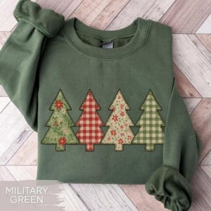 May include: Military green sweatshirt featuring four Christmas tree designs. The trees have floral and plaid patterns in red, green, and white. The sweatshirt has a crew neck and long sleeves.