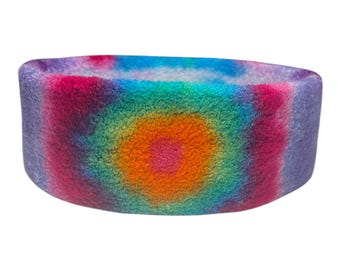 May include: A tie-dye headband in a range of colours. The design features concentric circles in shades of purple, pink, orange, green, and blue. The soft, plush material is visible in this close-up shot.